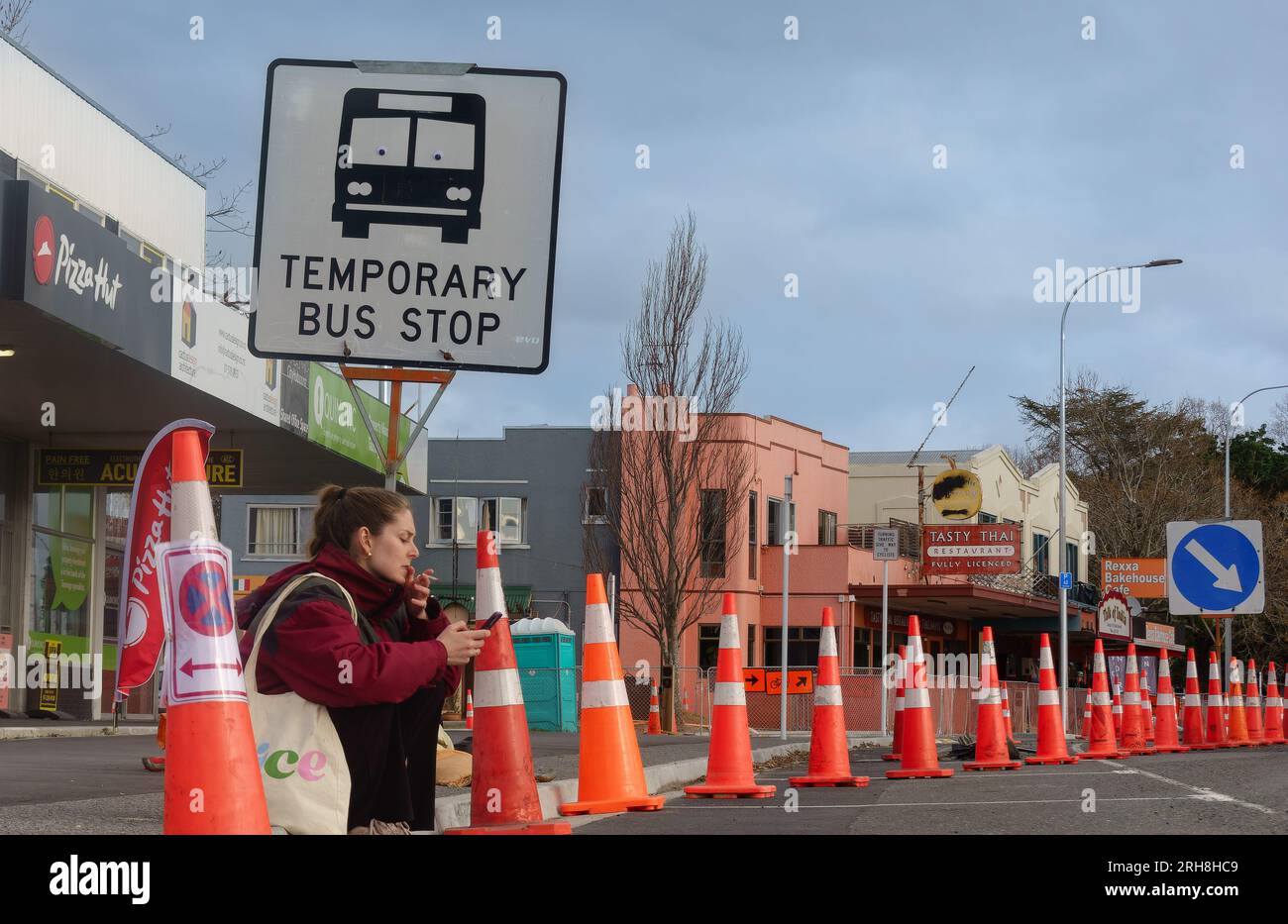 Tauranga Neuseeland - Juli 31 2023; Straßenkonen dominieren die Landschaft, während eine Frau auf dem Bordstein unter einem temporären Busschild sitzt und raucht und mobil aussieht Stockfoto