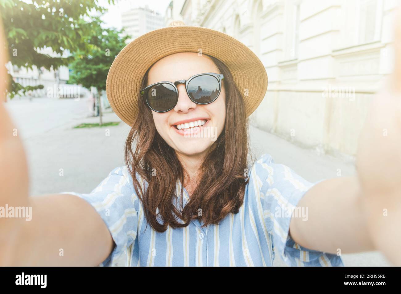 Junge lächelnde Frau mit Sonnenbrille und Hut, die auf der Straße steht und ein Selfie macht. Stockfoto