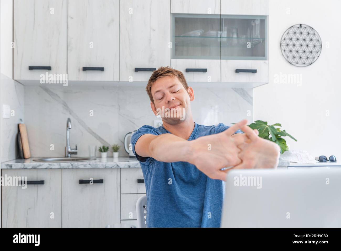 Junger, fröhlicher Mann, der sich die Hände streckt, während er zu Hause am Laptop arbeitet und Pause macht. Männlicher Freiberufler, der Computer für Fernarbeit nutzt Stockfoto