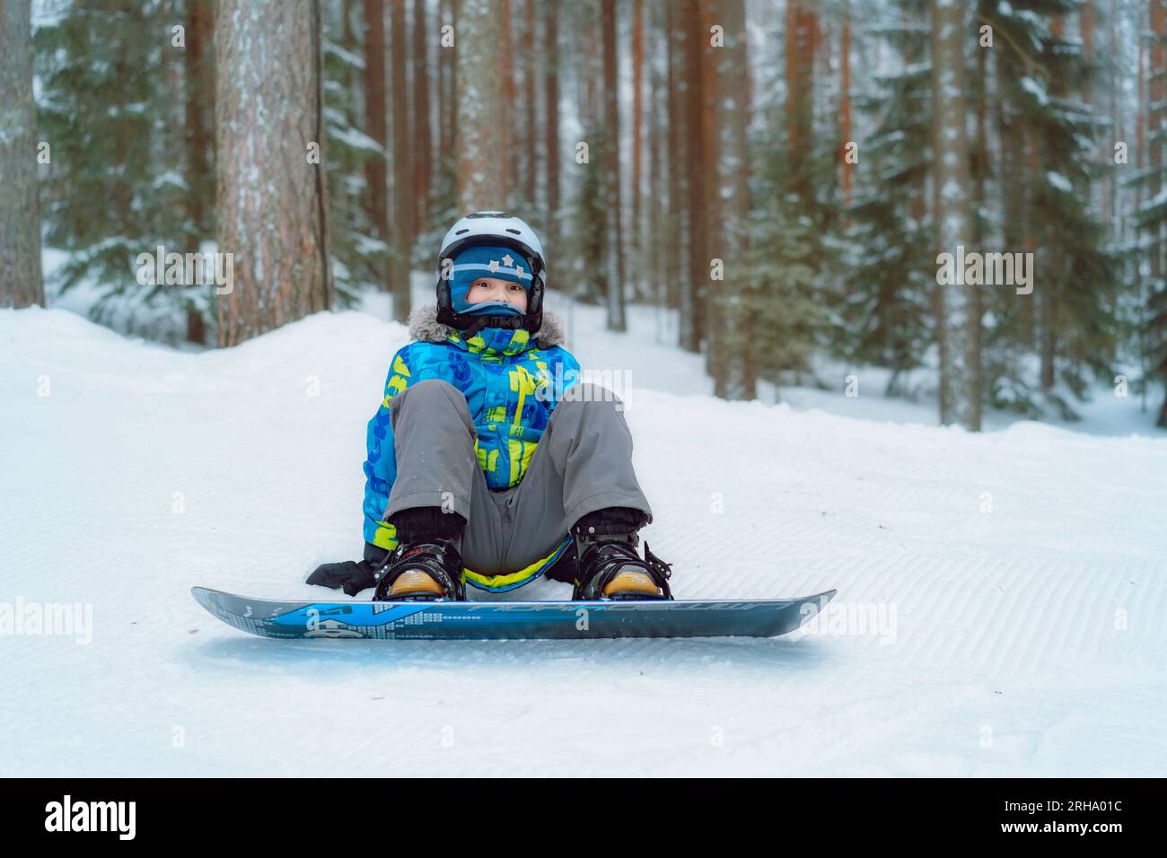 Der kleine Junge sitzt auf dem Schnee und legt seine Füße in Snowboardbindungen und stellt die Riemen ein. Bild mit selektivem Fokus. Hochwertiges Foto Stockfoto