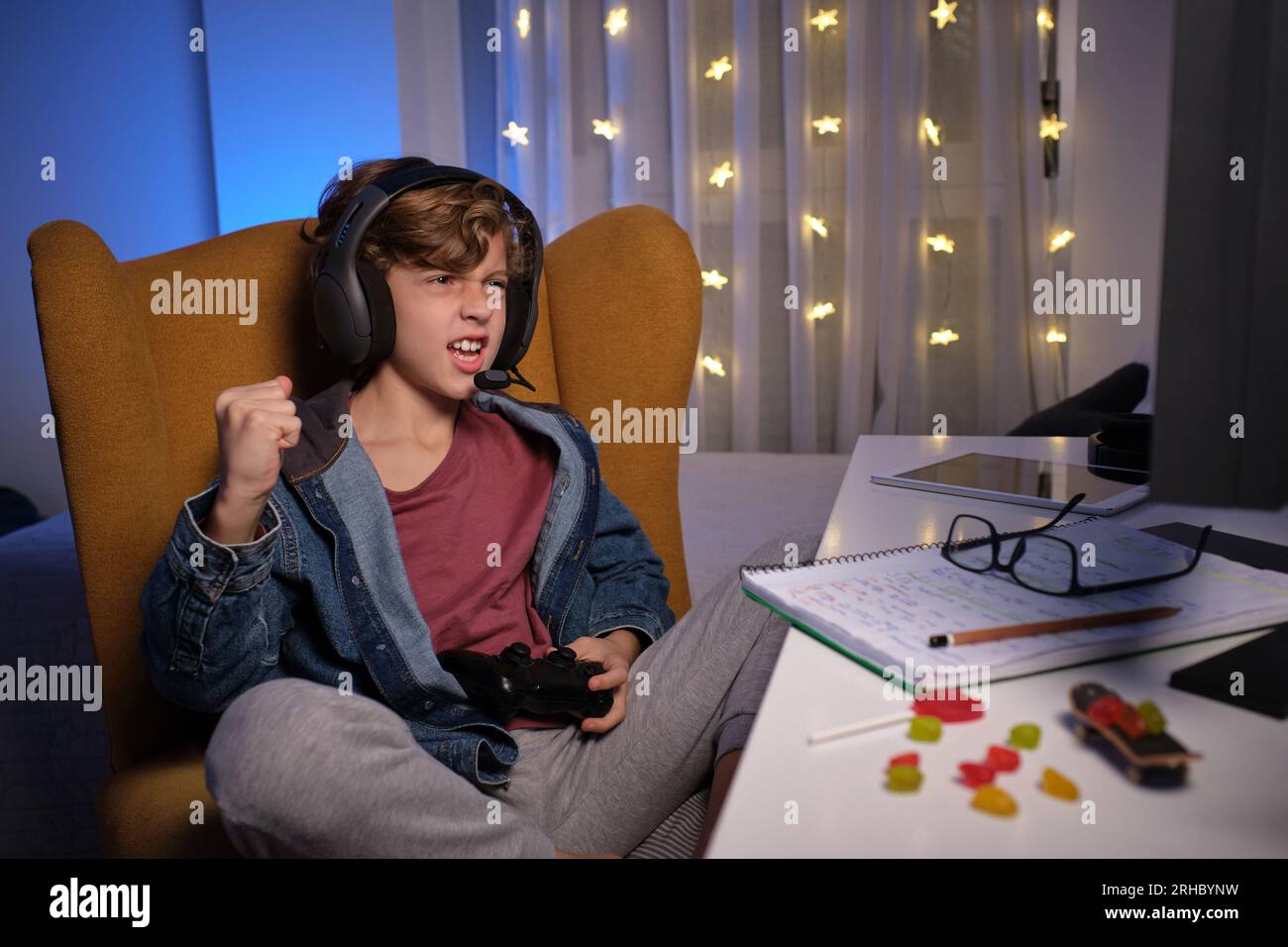 Ein Kind mit Kopfhörern knirscht sich die Faust, während es im Stuhl sitzt, und feiert den Erfolg des Videospiels bei schwachem Licht Stockfoto