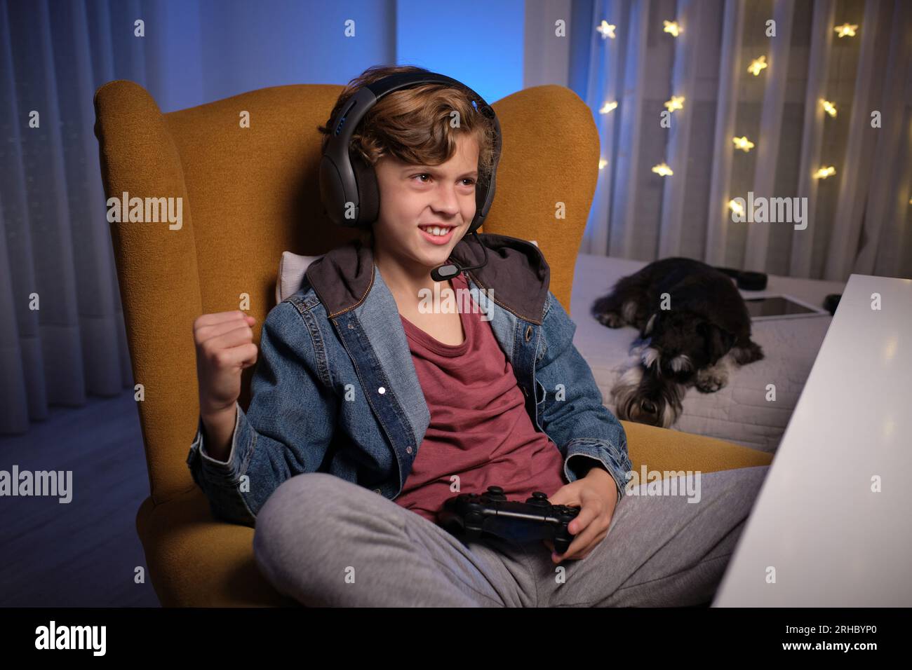 Positiver junger Junge im Headset sitzt auf einem bequemen Sessel mit Gamepad und feiert seine Leistung im Videospiel neben dem Bett mit dem Hund Stockfoto