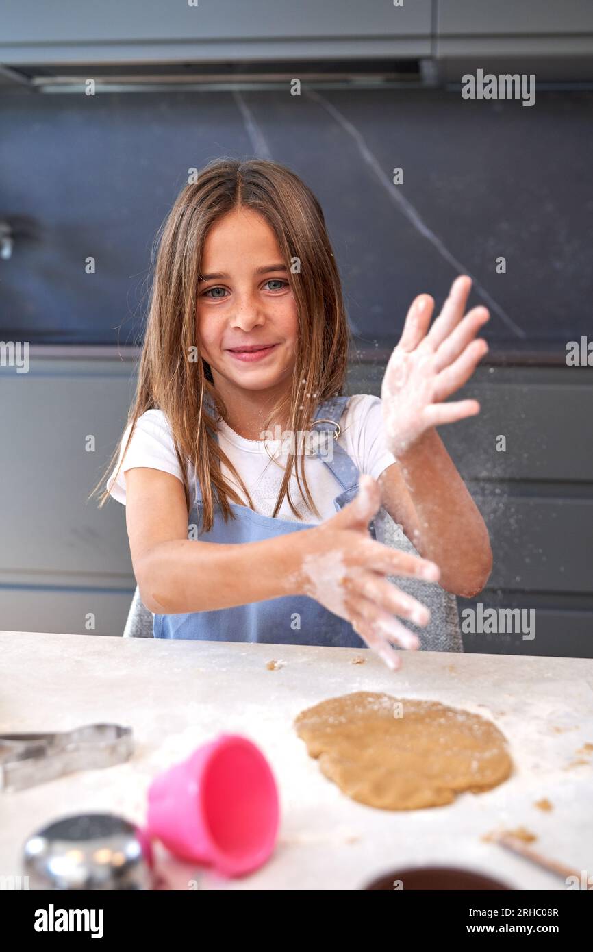 Entzückendes kleines Mädchen mit langen Haaren in der Schürze, das Mehl von den Händen schüttelt und nach dem Kneten von Teig während der Gebäckzubereitung in der Küche in die Kamera schaut Stockfoto