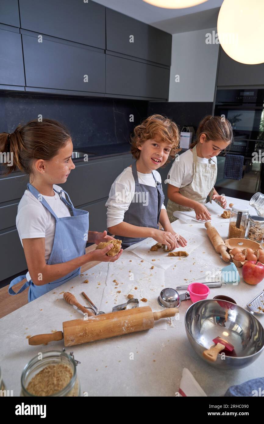 Gruppe von Kindern in Schürzen, die am Tisch in der modernen Küche stehen und Teig verarbeiten, während sie Kekse zubereiten Stockfoto
