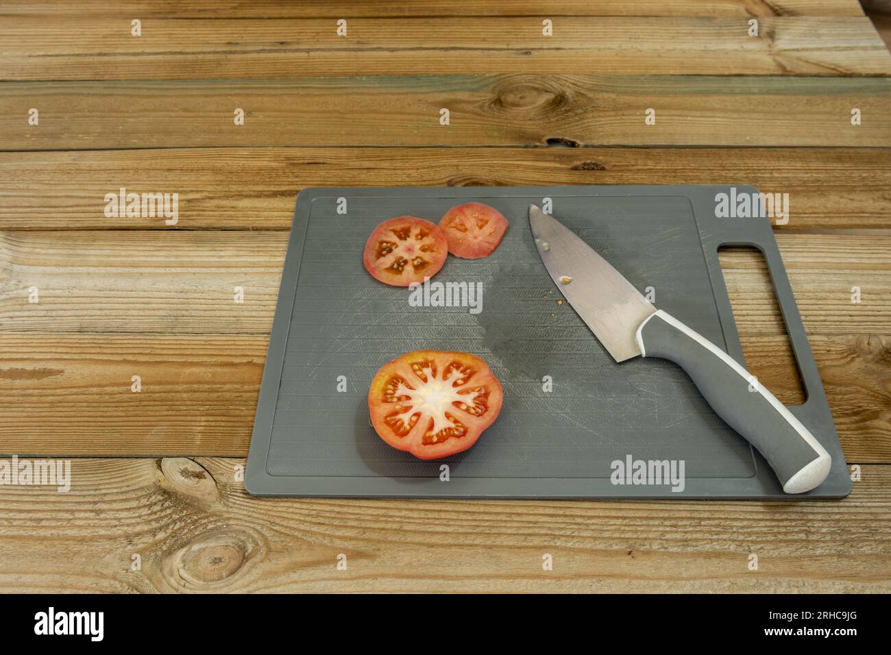Ein paar reife Tomatenscheiben, die auf einer grauen Kunststoffplatte mit einem scharfen Messer auf einem unlackierten Holztisch geschnitten werden Stockfoto