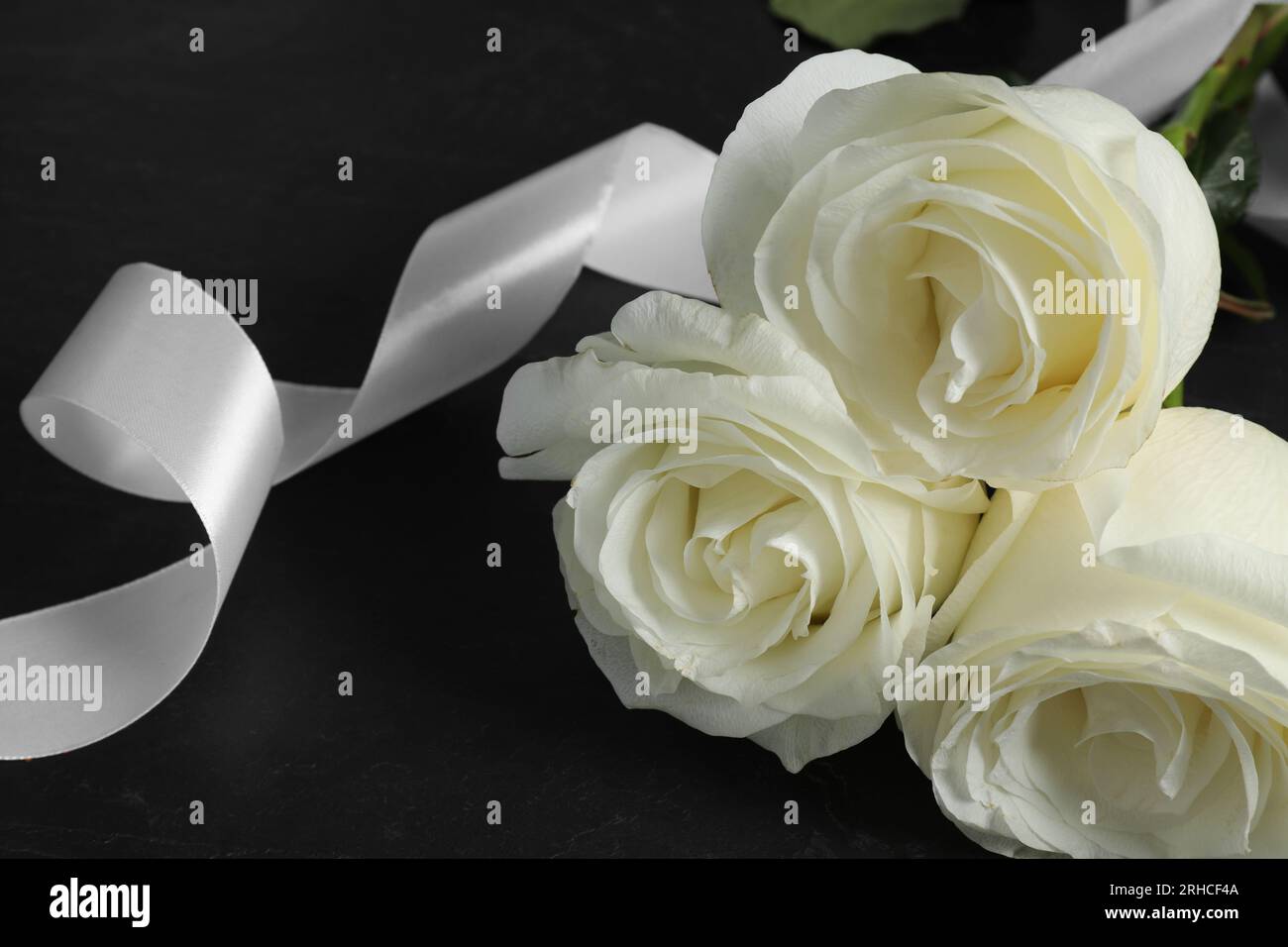 Weiße Rosen und Band auf schwarzem Tisch, Nahaufnahme. Beerdigungssymbole Stockfoto