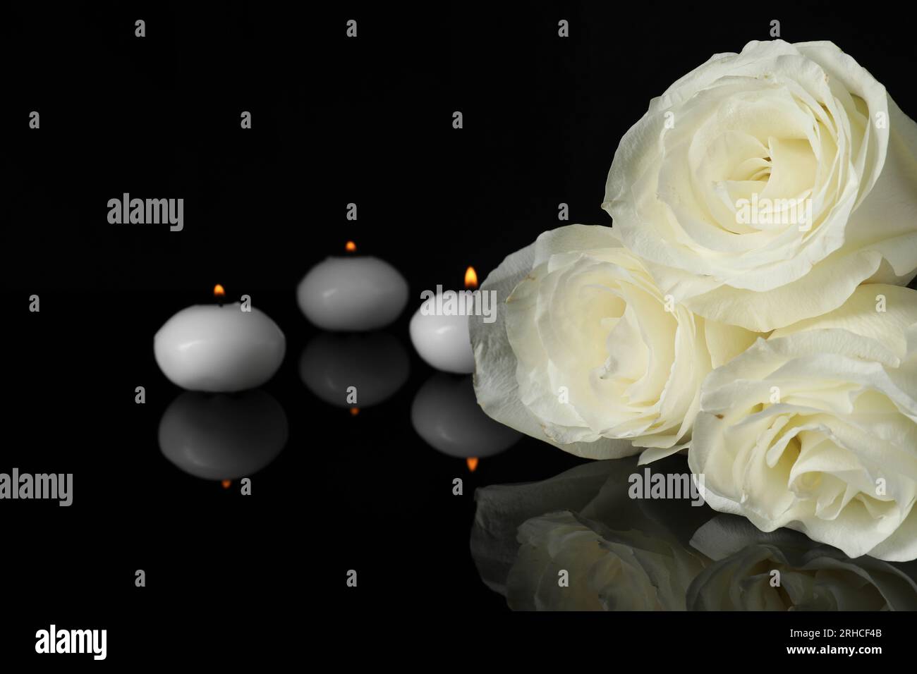 Weiße Rosen und brennende Kerzen auf schwarzer Spiegelfläche im Dunkeln, Nahaufnahme mit Platz für Text. Beerdigungssymbole Stockfoto