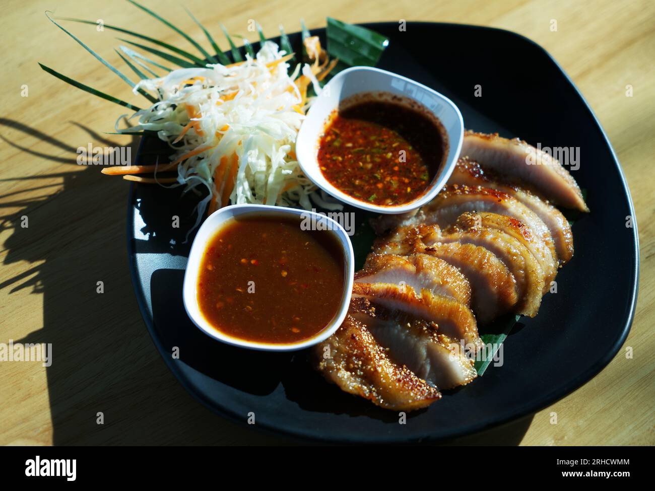 Thailändisches Essen gegrilltes Schweinehals oder Kor Moo Yang in thailändischer Sprache, geschnittenes Schweinefleisch mit zwei Arten Sauce und frischem Salat auf schwarzer Essensplatte auf Holzteller Stockfoto