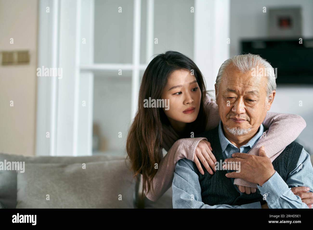 Trauriger älterer asiatischer Vater, der auf der Couch im Wohnzimmer zu Hause saß, getröstet von einer erwachsenen Tochter Stockfoto