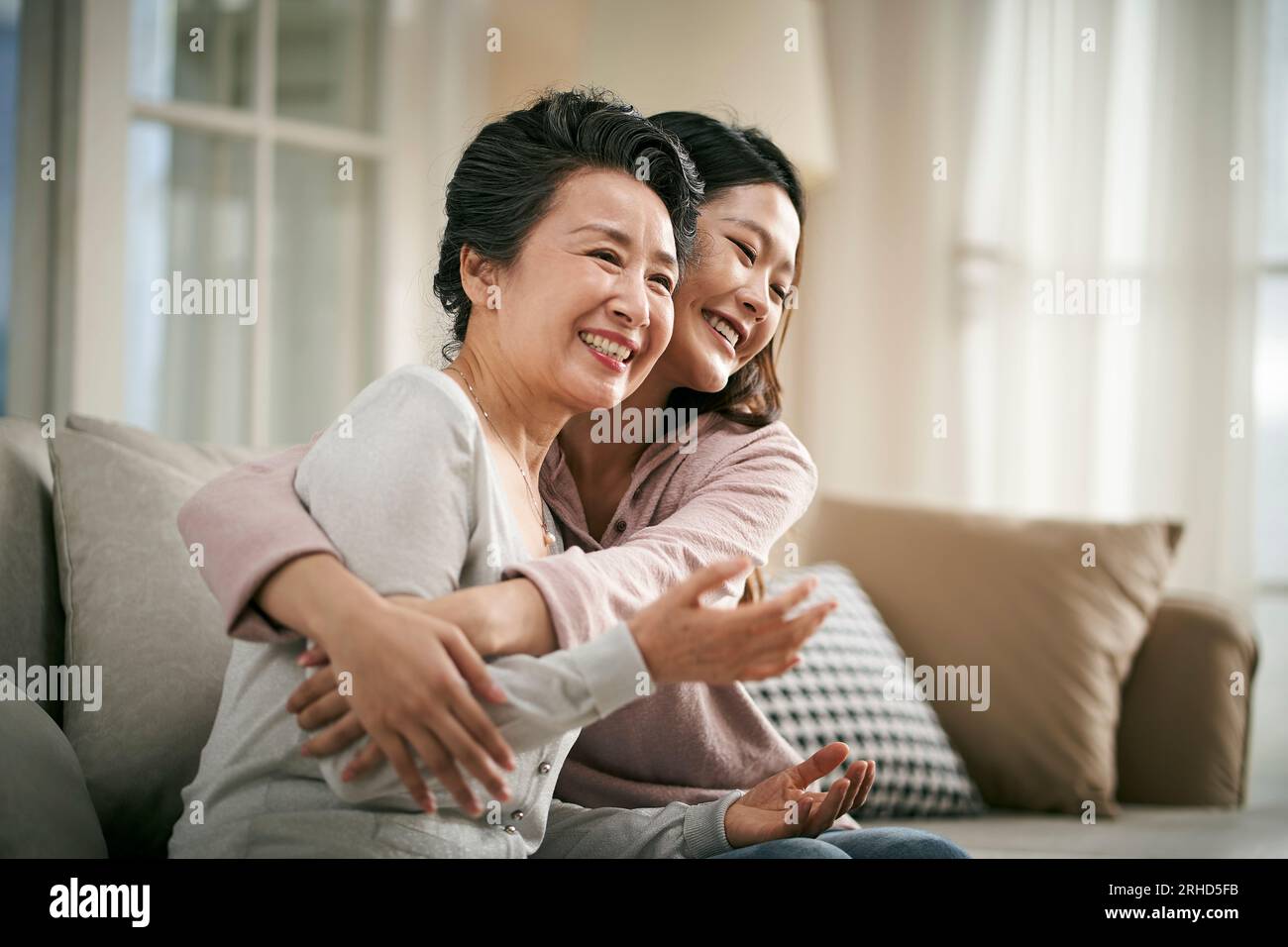 Liebende asiatische Erwachsene Tochter, die zu Hause auf der Couch sitzt und die ältere Mutter glücklich und lächelnd umarmt Stockfoto