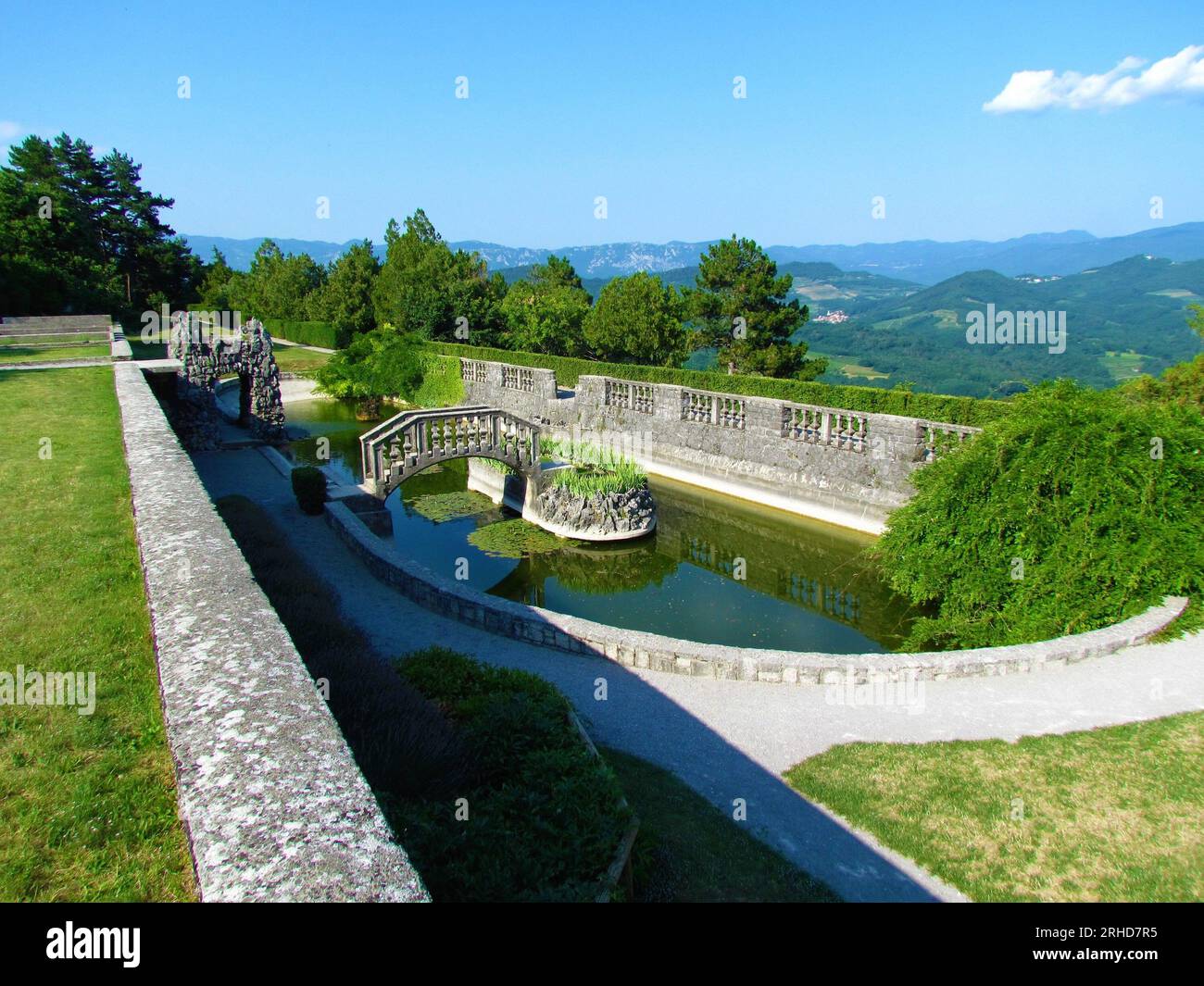 Ovaler Pool im Ferrari-Garten in Stanjel in der Littoral-Region von Slowenien mit einer Steinbrücke und einer kleinen Insel in der Mitte und einem Spiegelbild des bri Stockfoto