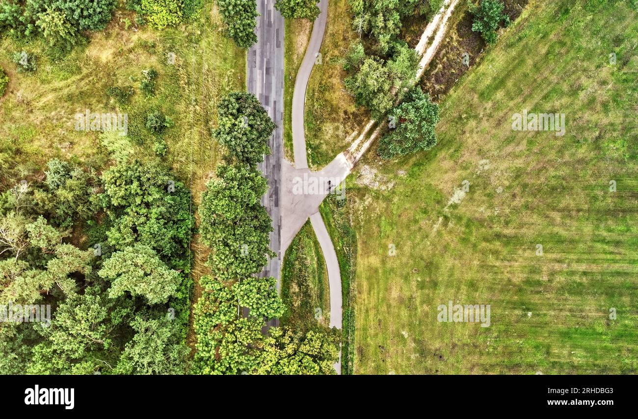 Vertikale abstrakte Luftaufnahme einer Landstraße und einer unbefestigten Straßenüberquerung mit einer Fahrradspur neben einer Wiese Stockfoto