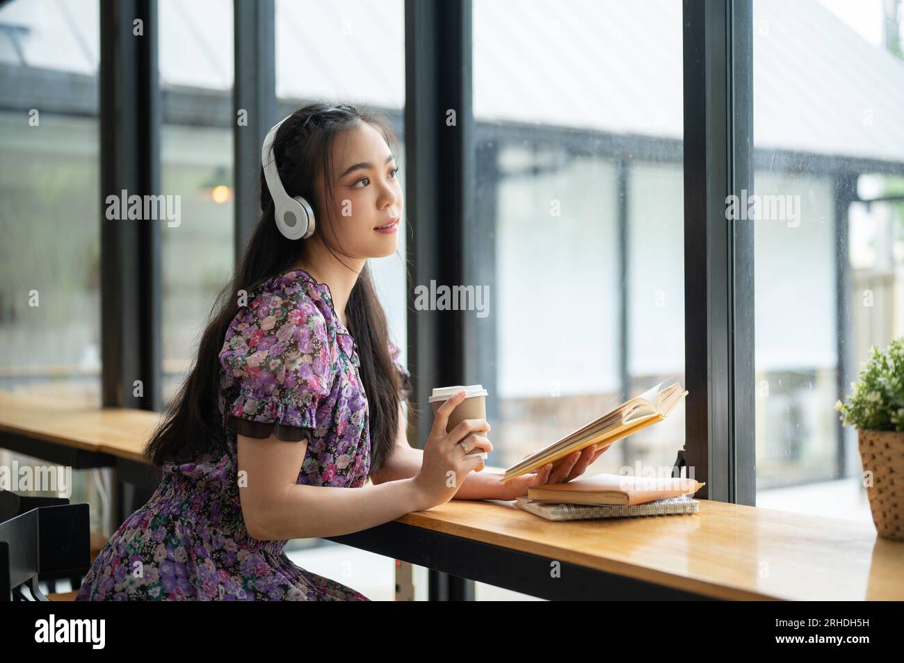 Eine schöne und charmante junge asiatische Frau in einem süßen Kleid träumt davon, während sie Musik auf ihren Kopfhörern hört und ein Buch in einem Café liest. Stockfoto
