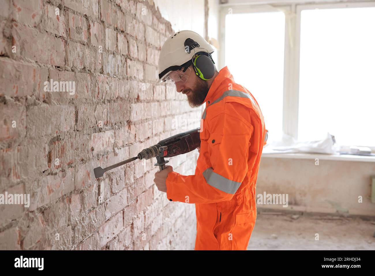 Bauarbeiter im orangefarbenen Overall, der alten Putz von der roten Ziegelwand mit einem Hammer entfernt. Stockfoto