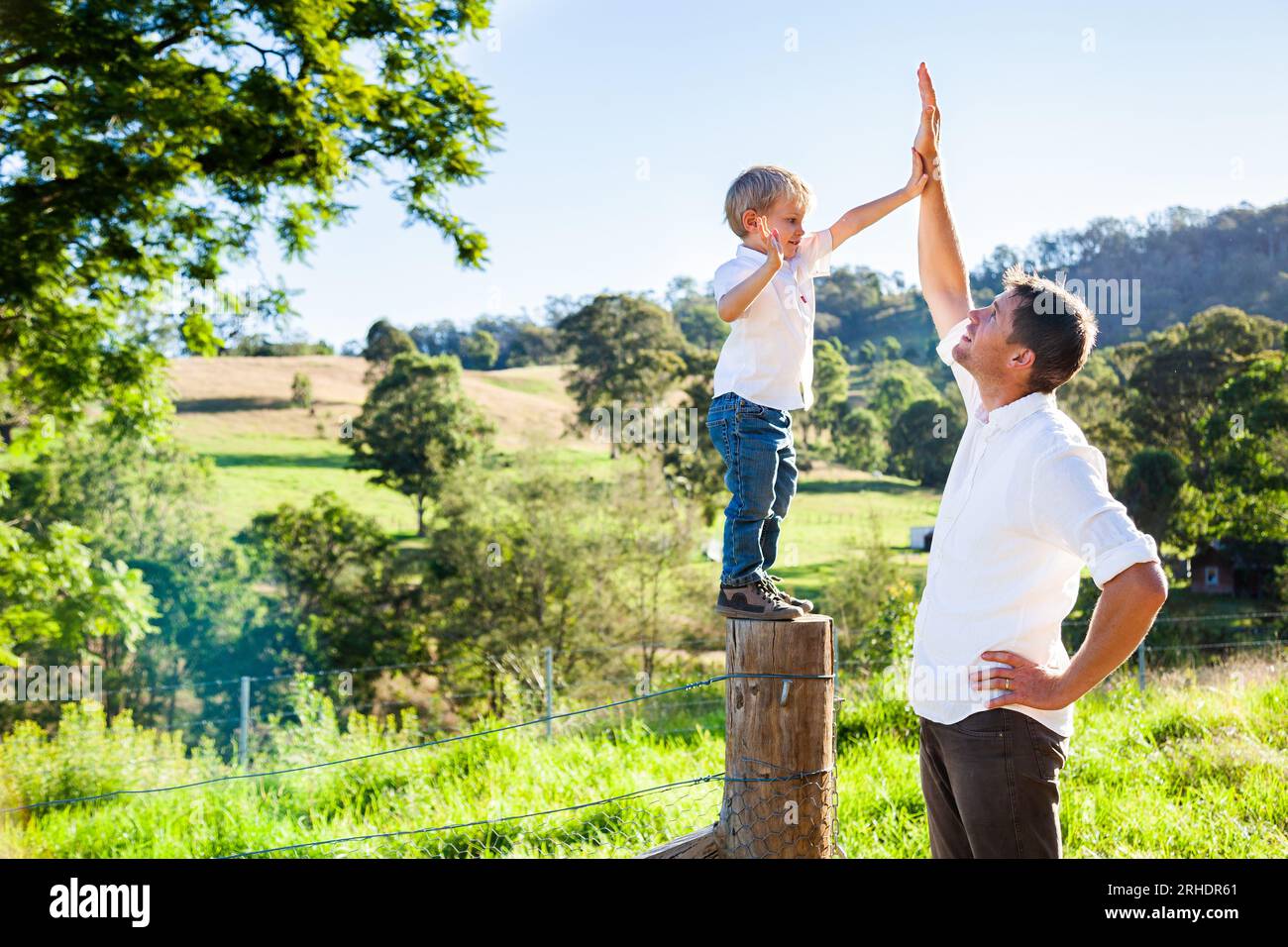 Vater und Sohn High Five auf dem sonnendurchfluteten Bauernhof, der auf einem Zaunsiebpfosten steht Stockfoto