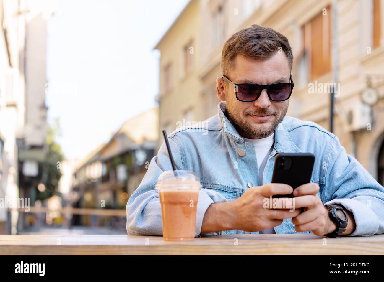 Junger Erwachsener, der eine Sonnenbrille und eine Jeansjacke trägt und sein Smartphone benutzt, während er sich im Straßencafé in der Stadt entspannt. Stockfoto