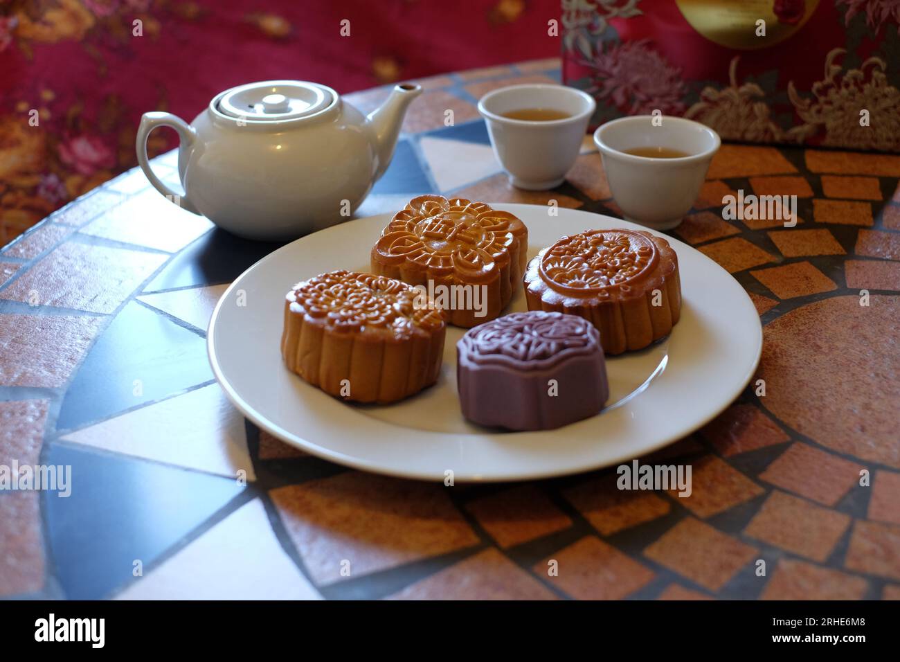 Vier verschiedene chinesische Mondkuchen auf einem weißen Teller, rundes, quadratisches Gebäck und ein violetter sechseckiger Kuchen, zwei Teetassen und eine Teekanne. Stockfoto