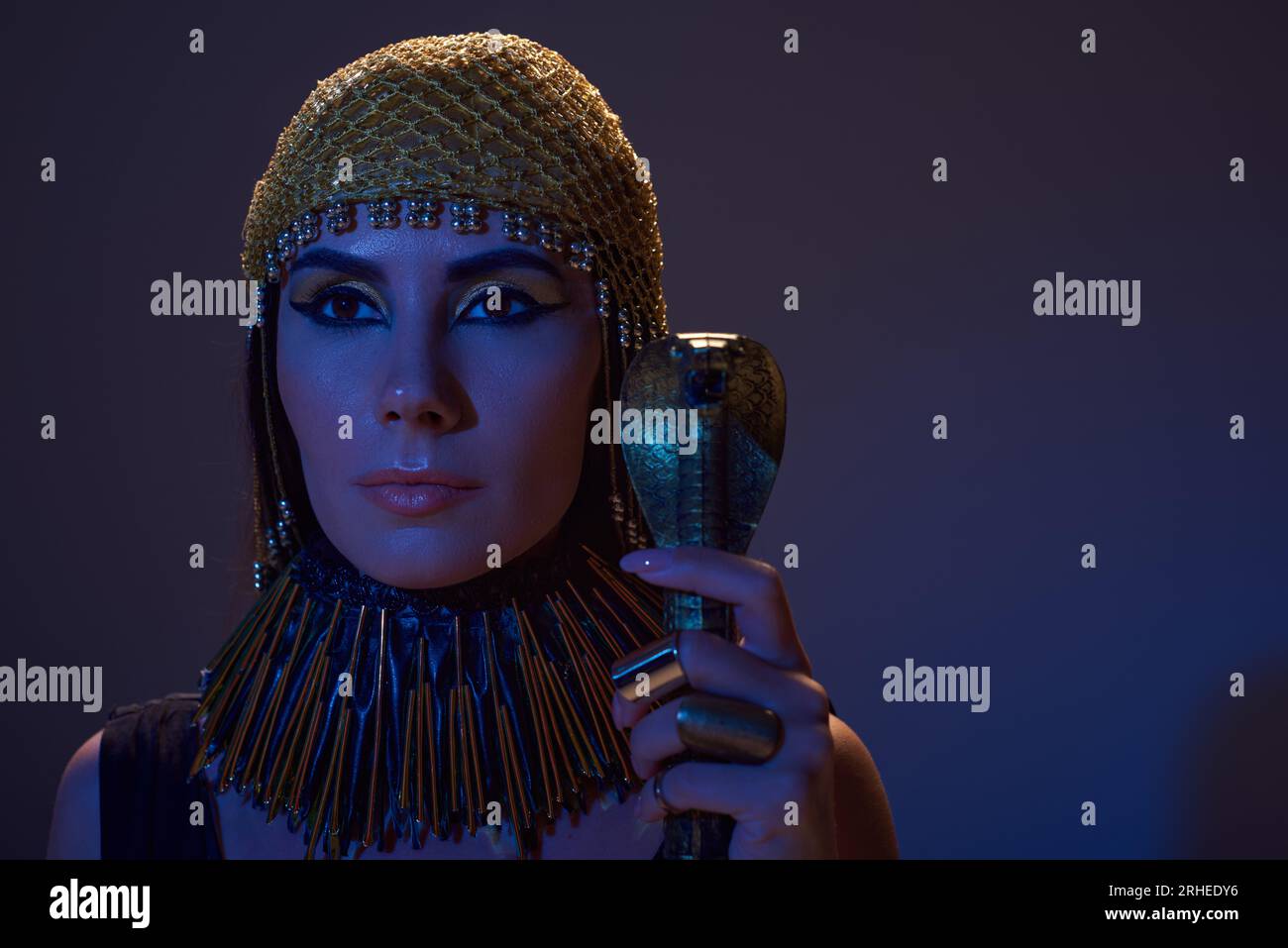 Eine Frau in ägyptischem Kopfschmuck und Make-up, die einen Gauner in Schlangenform auf blauem Licht auf Braun hält Stockfoto