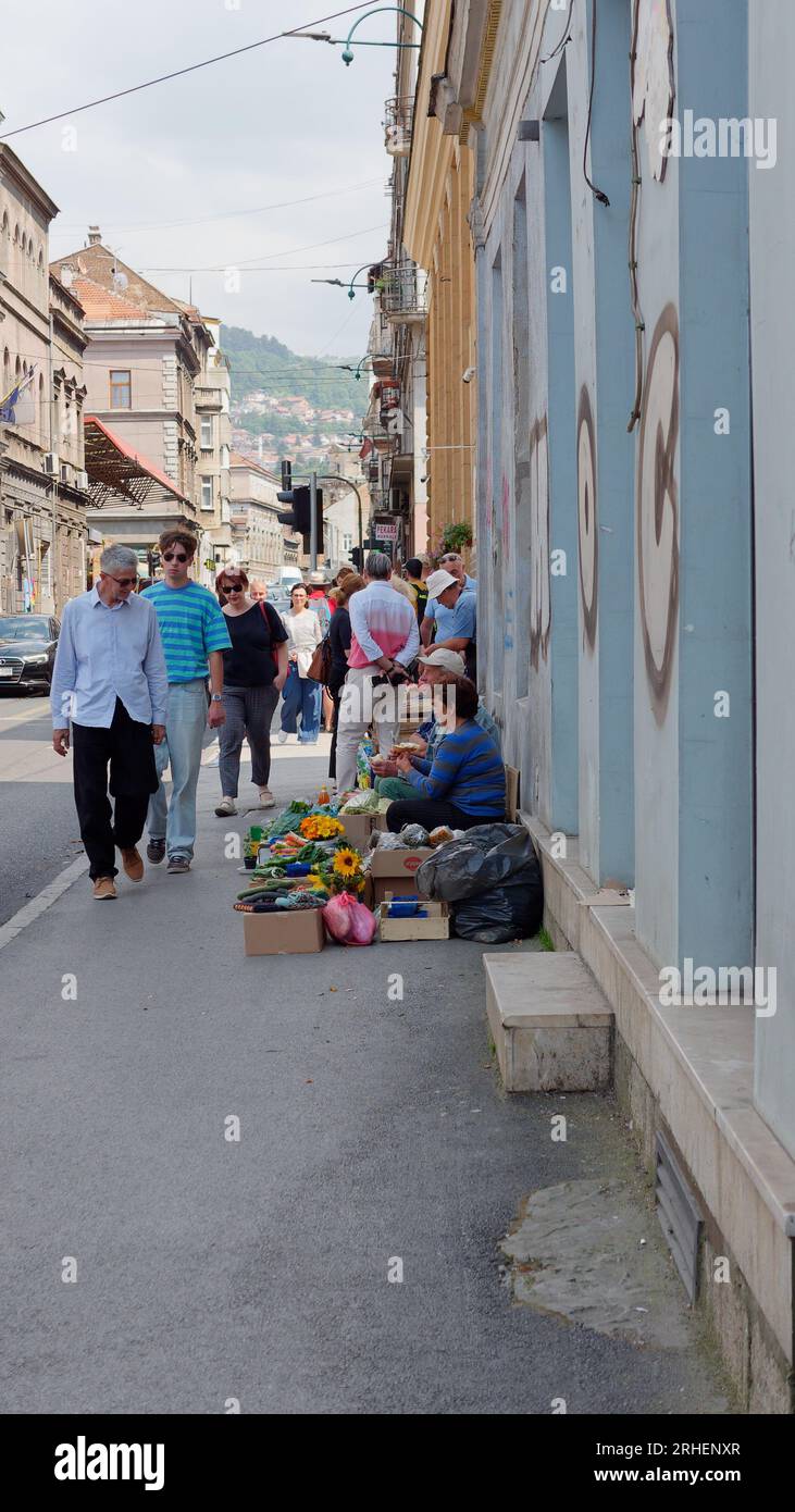 Einheimische sitzen auf dem Bürgersteig und verkaufen Blumen, Obst und Gemüse, während Passanten durch die Waren stöbern. Sarajevo, Bosnien und Herzegowina, 16. August 2023. Stockfoto