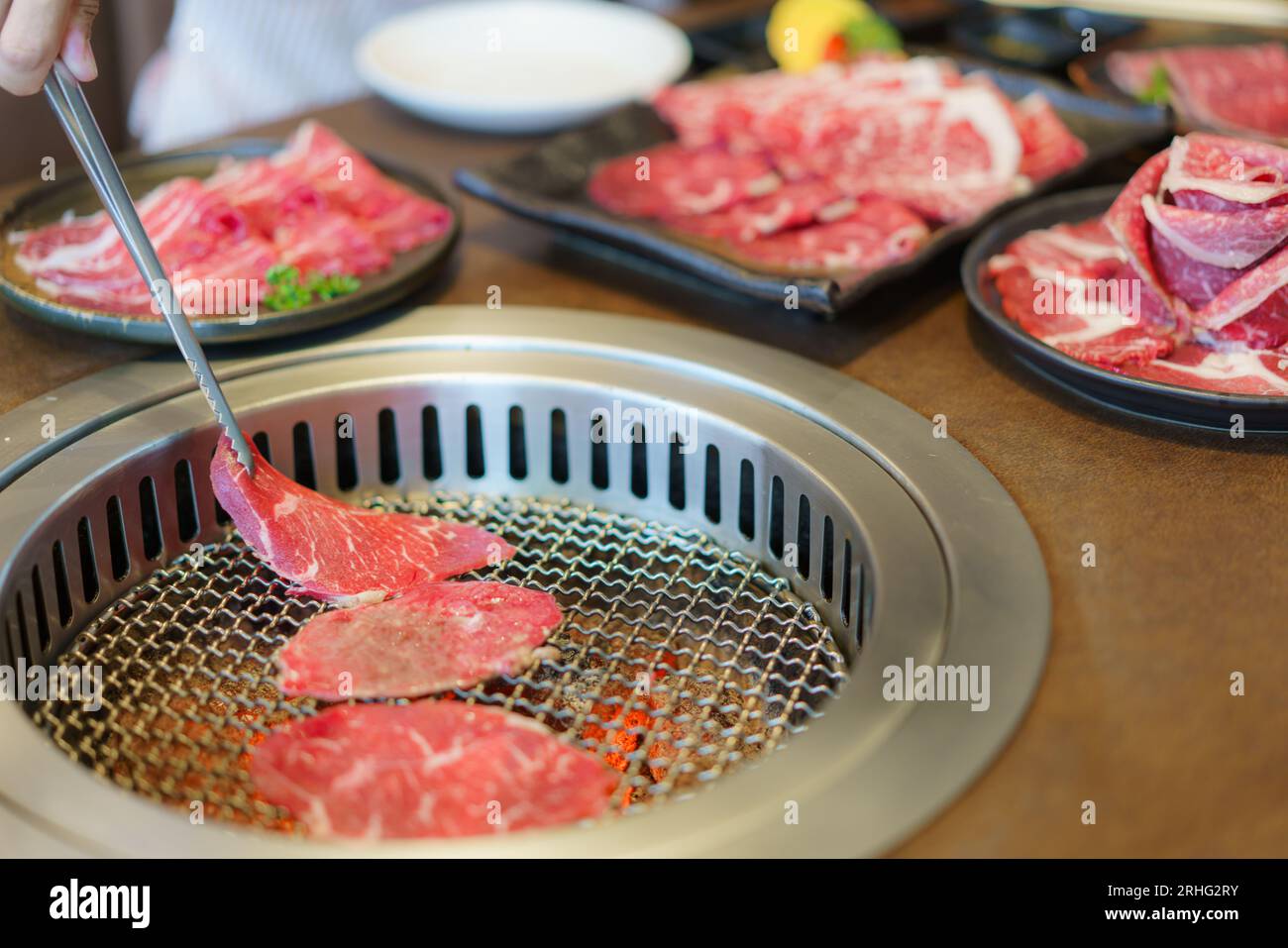Die Hand einer Frau verwendet Zangen, um Wagyu-Rindfleisch auf einen Teller zu legen, bereit zum Grillen über Holzkohle, was das kulinarische Erlebnis in einem japanischen Restaurant bereichert Stockfoto