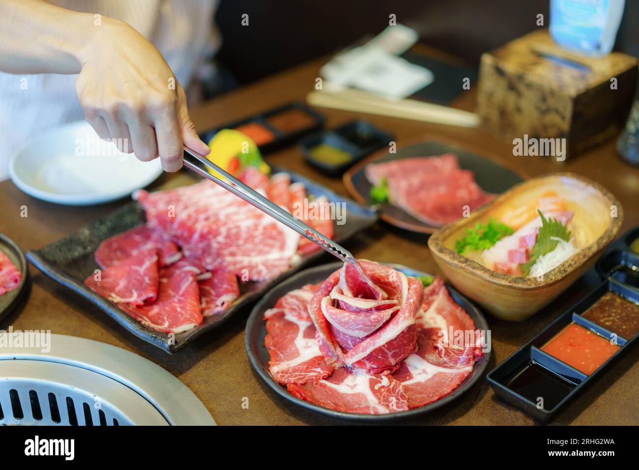 Die Hand einer Frau verwendet Zangen, um Wagyu-Rindfleisch auf einen Teller zu legen, bereit zum Grillen über Holzkohle, was das kulinarische Erlebnis in einem japanischen Restaurant bereichert Stockfoto