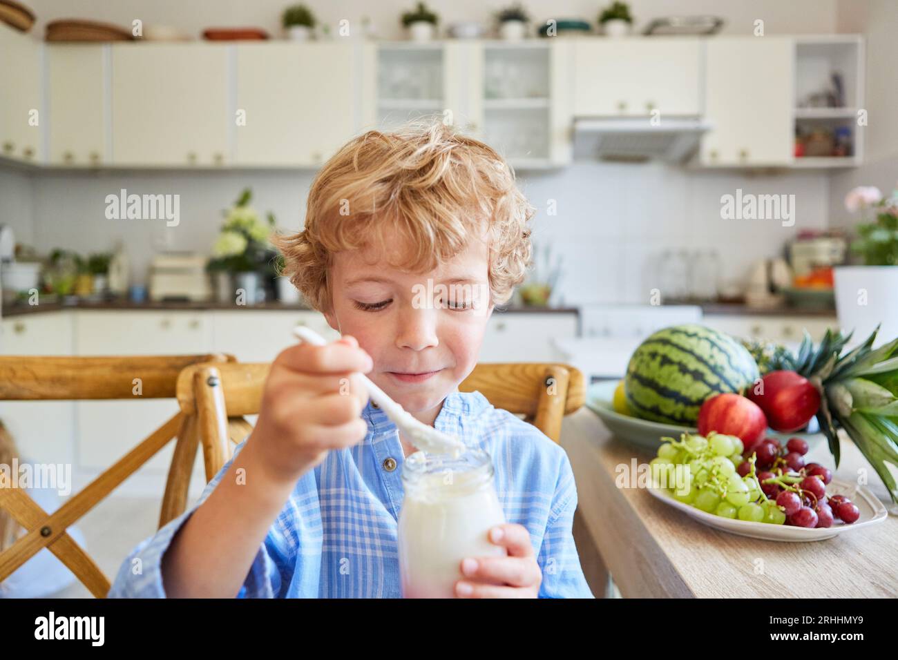 Niedlicher blonder Junge, der frisches Joghurtparfait isst, während er in der Küche zu Hause neben dem Tisch sitzt und Früchte trägt Stockfoto