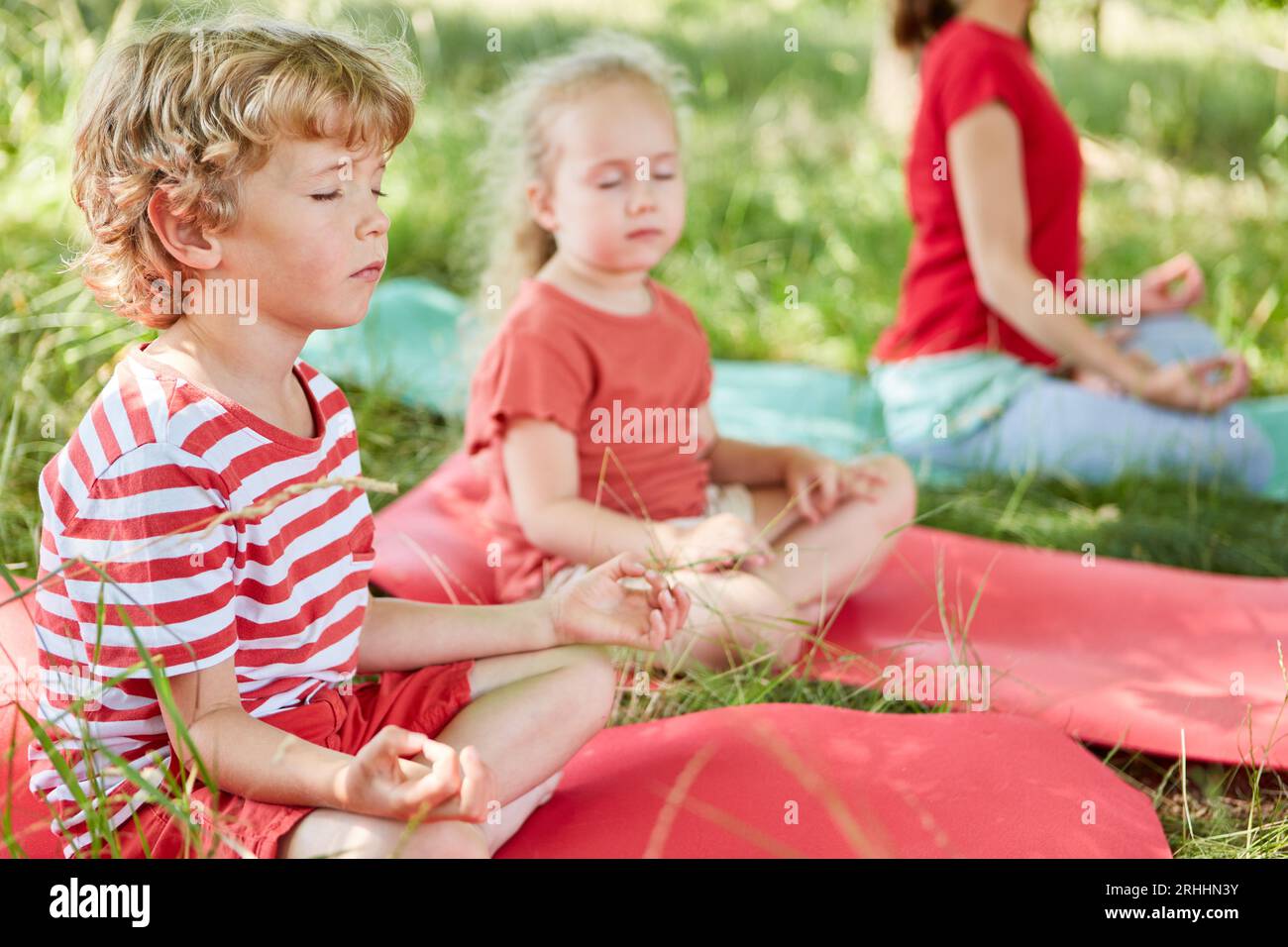 Junge mit geschlossenen Augen meditierend, während er auf der Matte neben der Schwester im Park sitzt Stockfoto