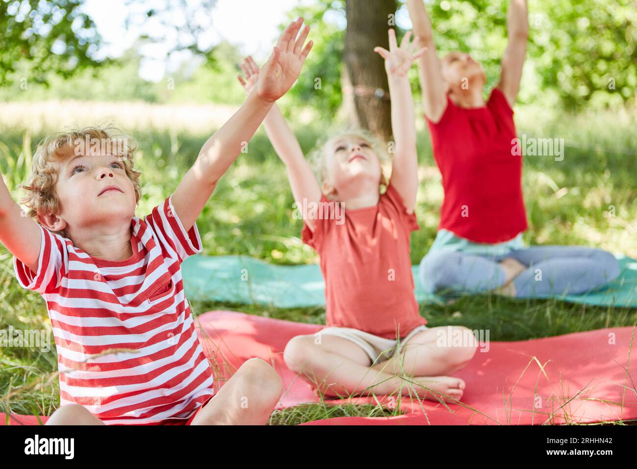 Die Familie sitzt auf einer Matte mit erhobenen Armen, während sie im Park Sport treibt Stockfoto