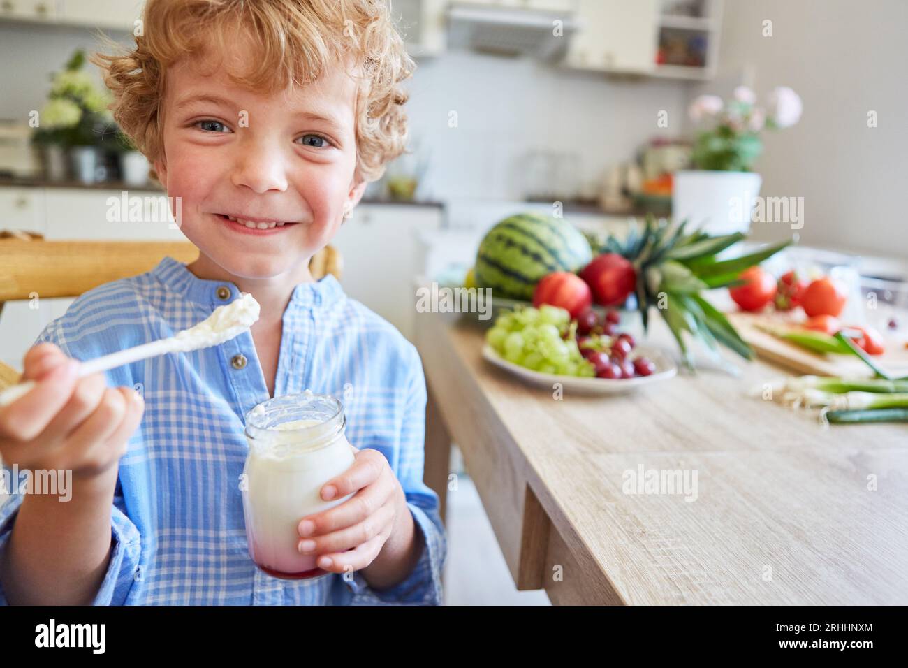 Porträt eines lächelnden blonden Jungen, der ein Glas frischen Parfait hält, während er zu Hause in der Küche sitzt Stockfoto