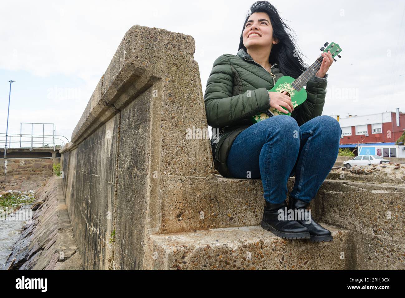 Joven mujer venezolana sonriendo muy feliz disfrutando el fin de semana con su ukulele, tocando sentada en las escaleras junto al rio en buenos aires. Stockfoto