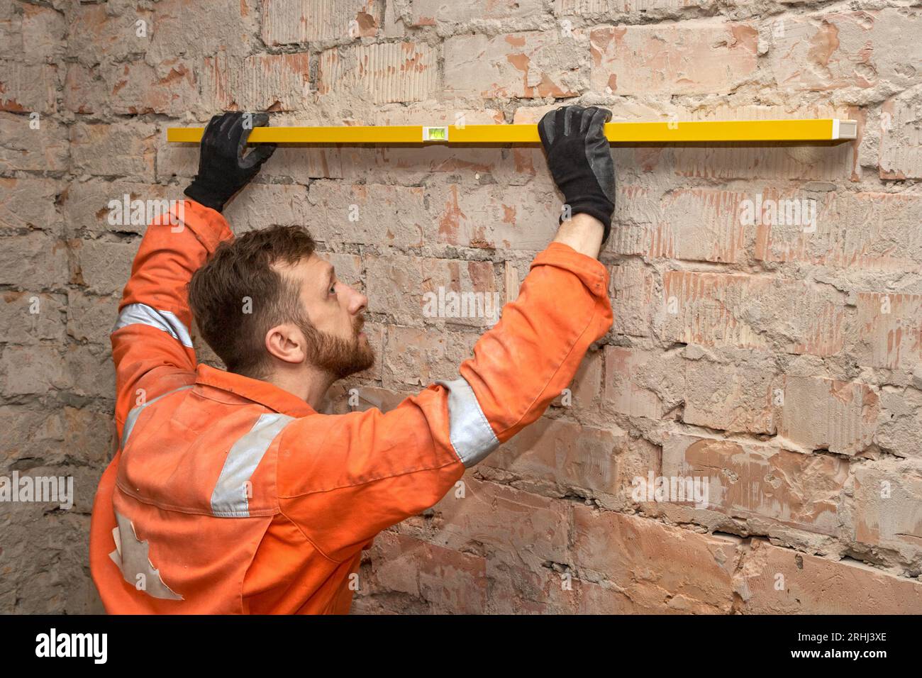 Junger Bauarbeiter, der die Ziegelwand auf Ebene überprüft, Handschuhe und orangefarbene Overalls trägt. Stockfoto