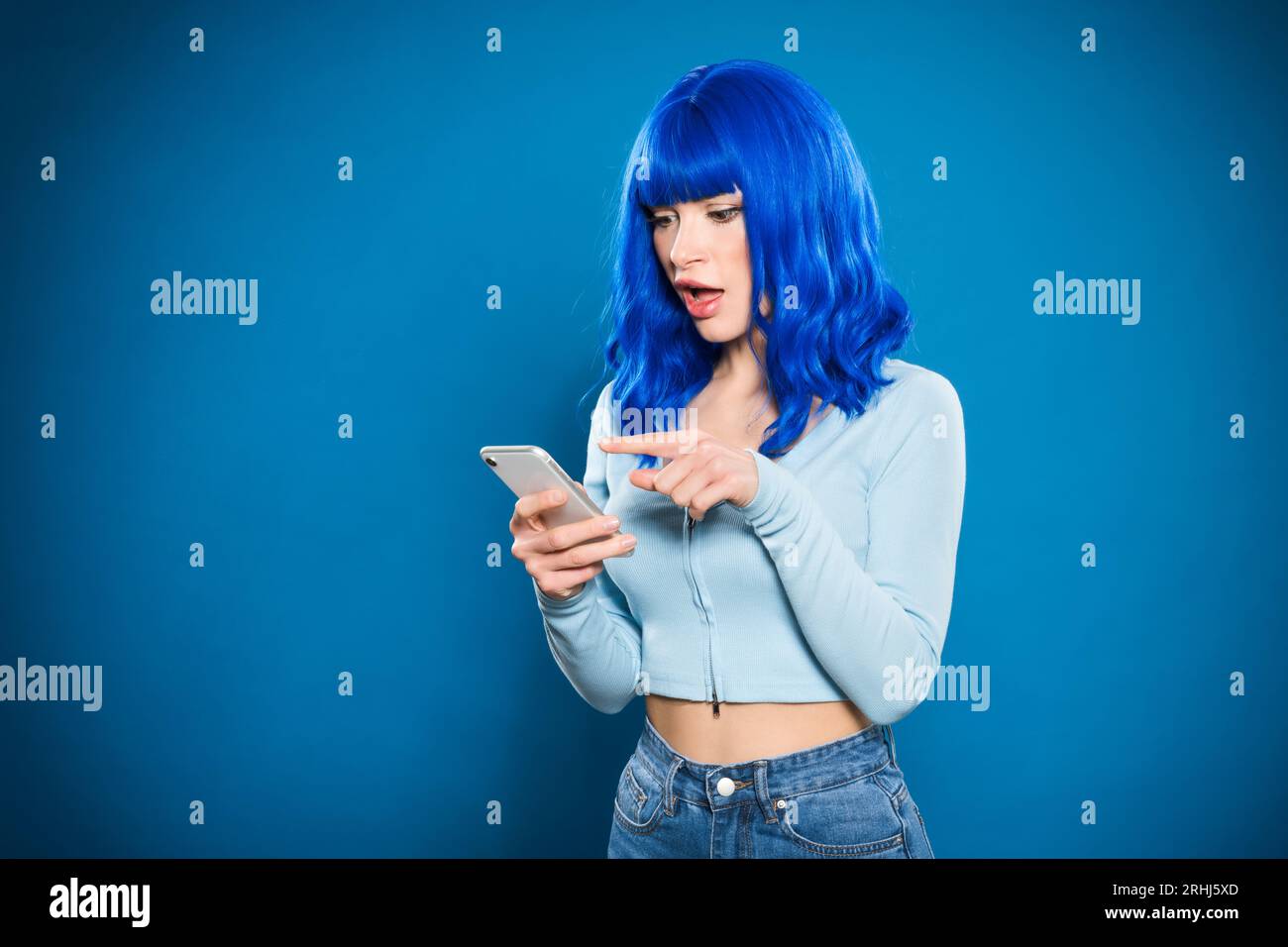 Staunende junge Frau mit blauem Haar, die auf den Bildschirm des Mobiltelefons schaut und mit dem Finger vor einem leuchtend beleuchteten Hintergrund steht Stockfoto