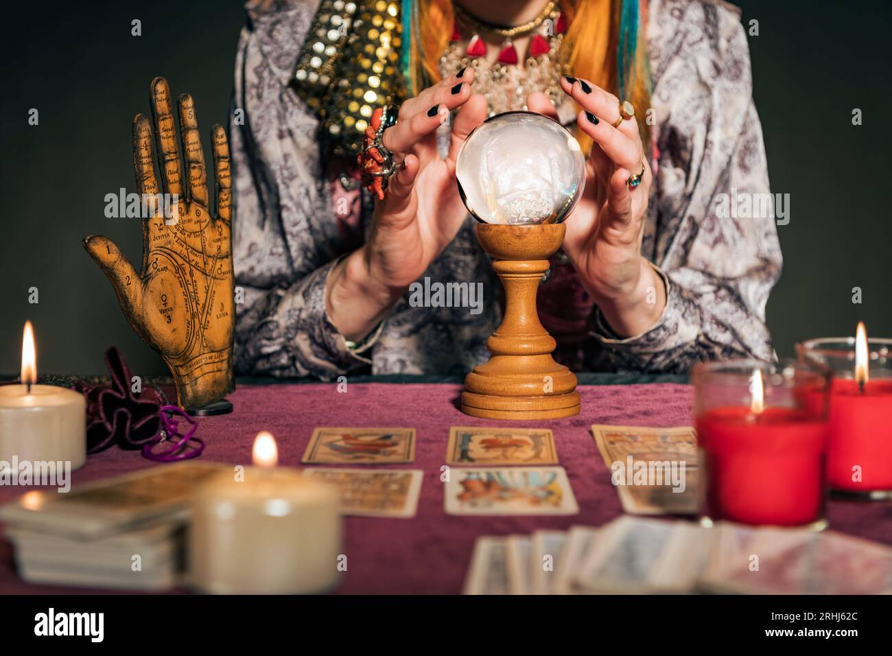 Baue Wahrsagerin in authentischer Kleidung, die mit brennenden Kerzen und Tarotkarten am Tisch sitzt, während du mit dem Zauberball die Zukunft erraten kannst Stockfoto