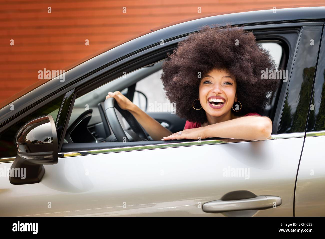 Lächelnde junge afroamerikanische Frau mit Afro-Frisur, die auf die Kamera schaut, während sie im geparkten Auto auf dem Fahrersitz sitzt, mit der Hand auf dem Lenkrad nea Stockfoto