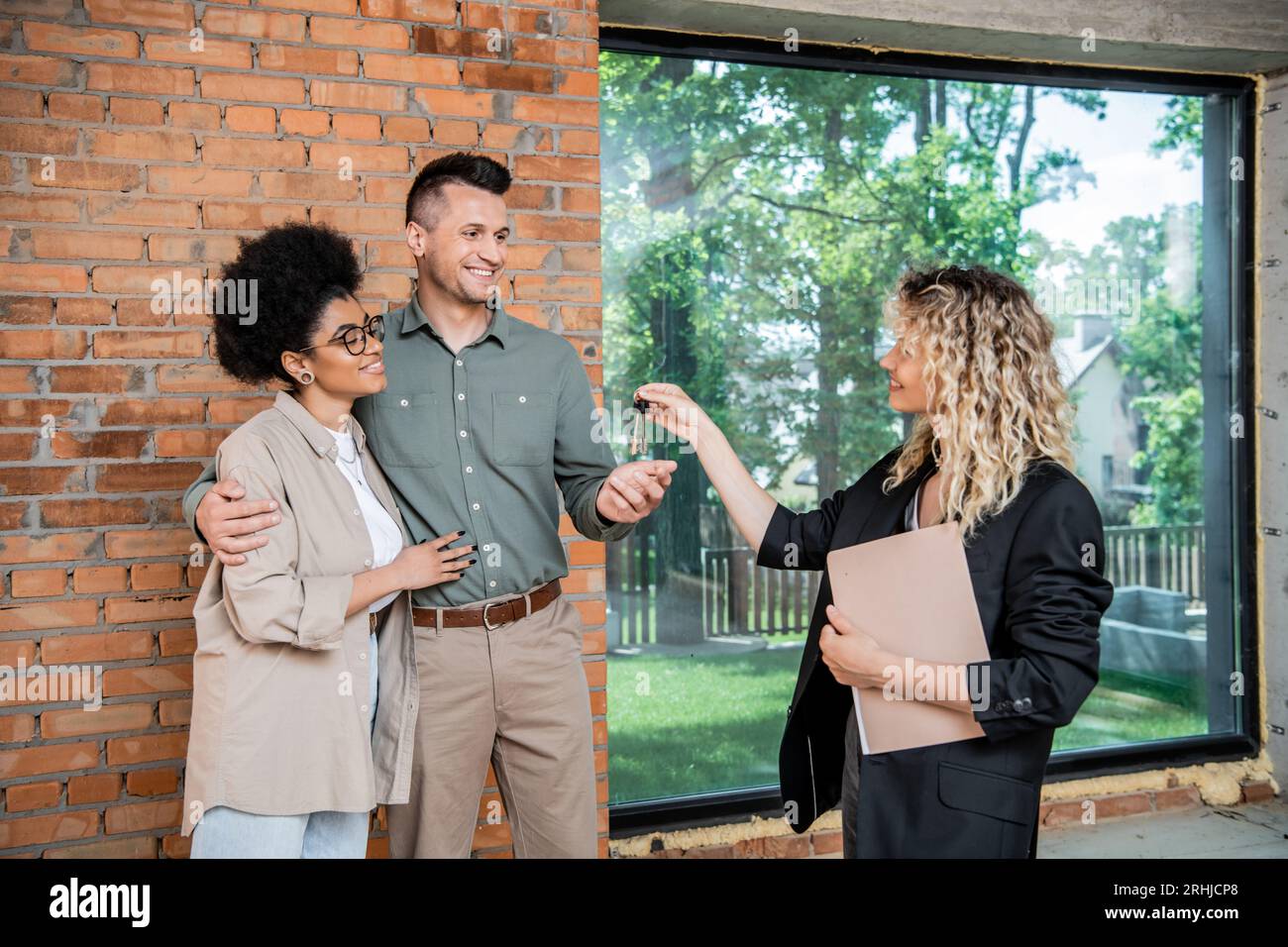 Immobilienmakler mit Dokumenten, die Schlüssel aus dem neuen Haus an zufriedene multikulturelle Paare geben Stockfoto