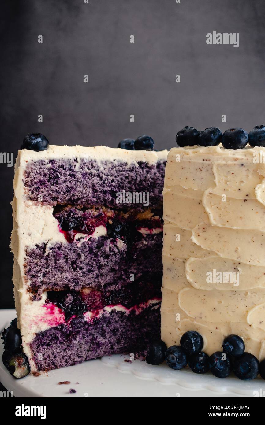 Eine Scheibe Blaubeerkobbler-Kuchen entfernen: Dreischichtiger, in Scheiben geschnittener Kuchen mit Blaubeerstreuselfüllung und Vanillebuttercreme-Zuckerguss Stockfoto