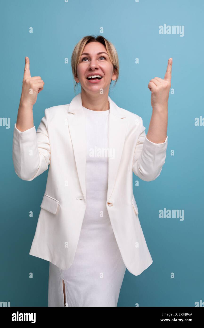 Fröhliche, hübsche, blonde, junge europäische Frau in einer weißen Jacke und Kleid zeigt mit dem Finger auf die Werbung Stockfoto
