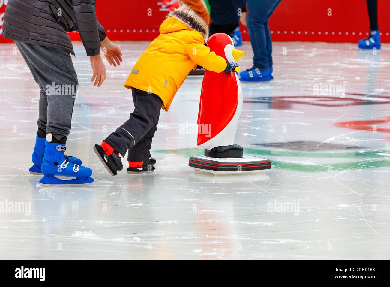 Ein Kind, begleitet von einem Erwachsenen, lernt auf einer Eislaufbahn Schlittschuh zu laufen und hält an einem Assistenten, einem Spielzeugpinguin, fest. Stockfoto