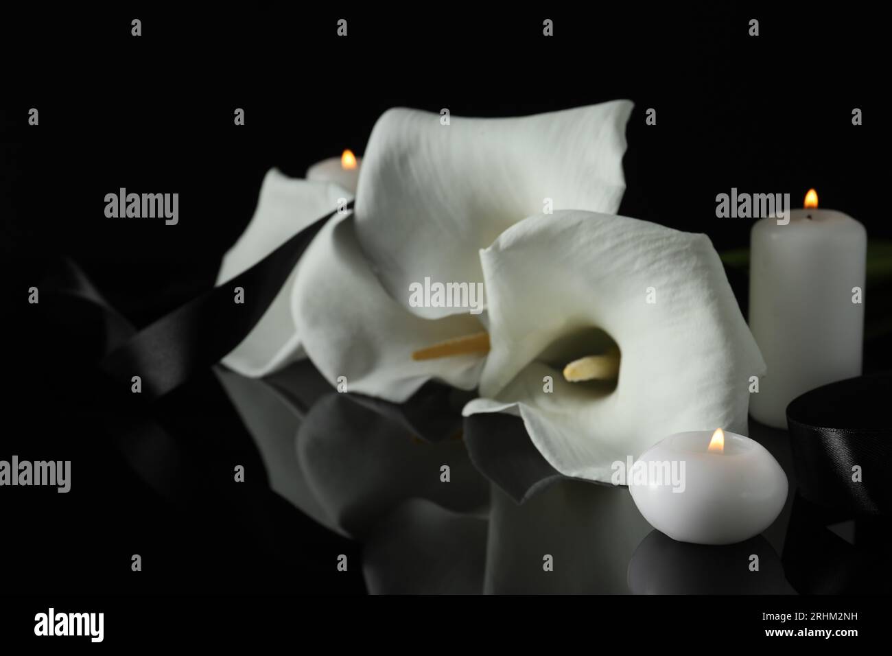 Brennende Kerzen, Band und weiße Calla-Lilie-Blüten auf schwarzer Spiegeloberfläche in Dunkelheit, Nahaufnahme. Beerdigungssymbole Stockfoto