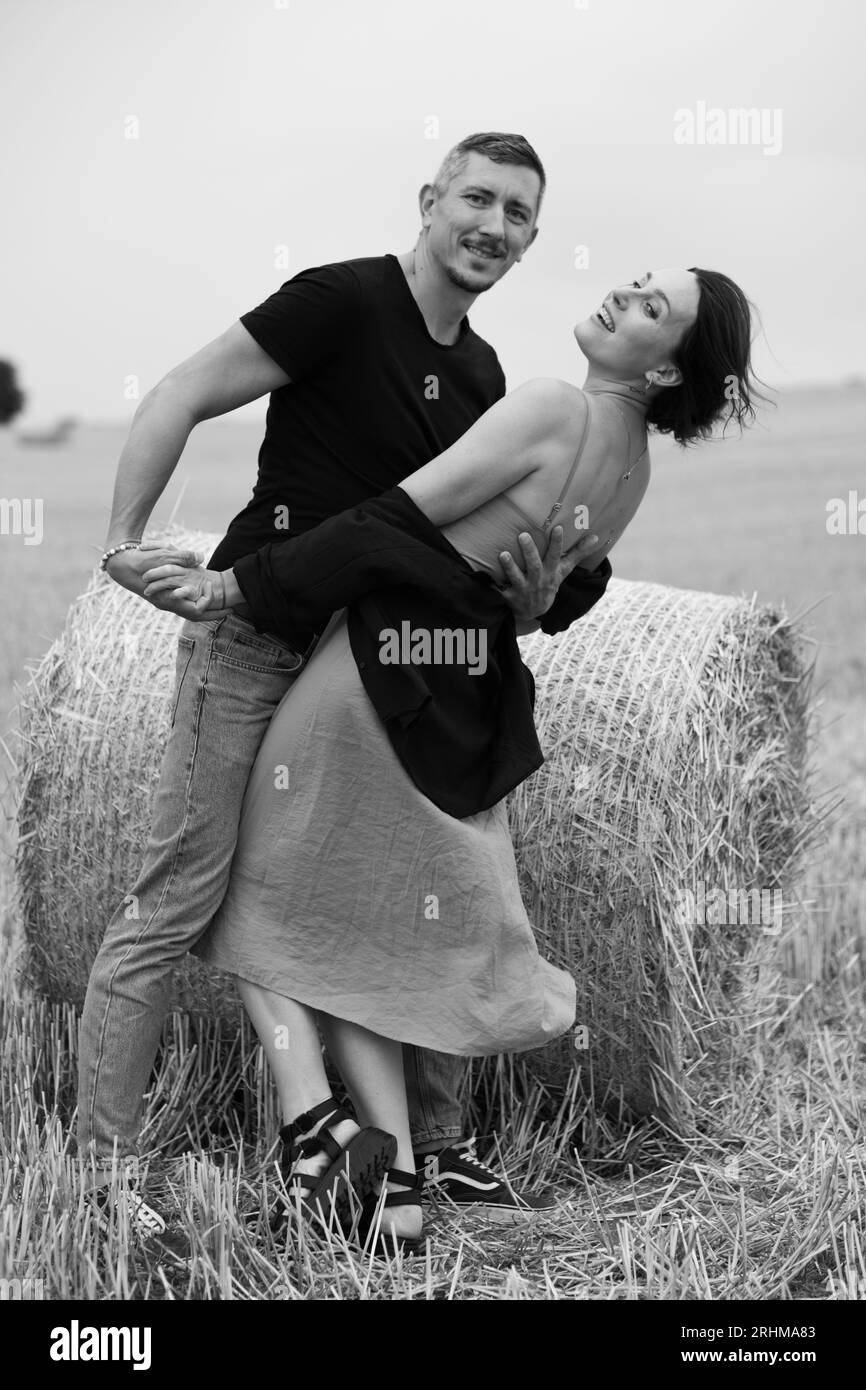 Ein junges Paar wunderschöner Leute hat Spaß auf dem Feld in der Nähe von runden Heuballen. Ein Mann, der mit seiner Frau in der Nähe eines Strohballens tanzt. Schwarz und weiß Stockfoto
