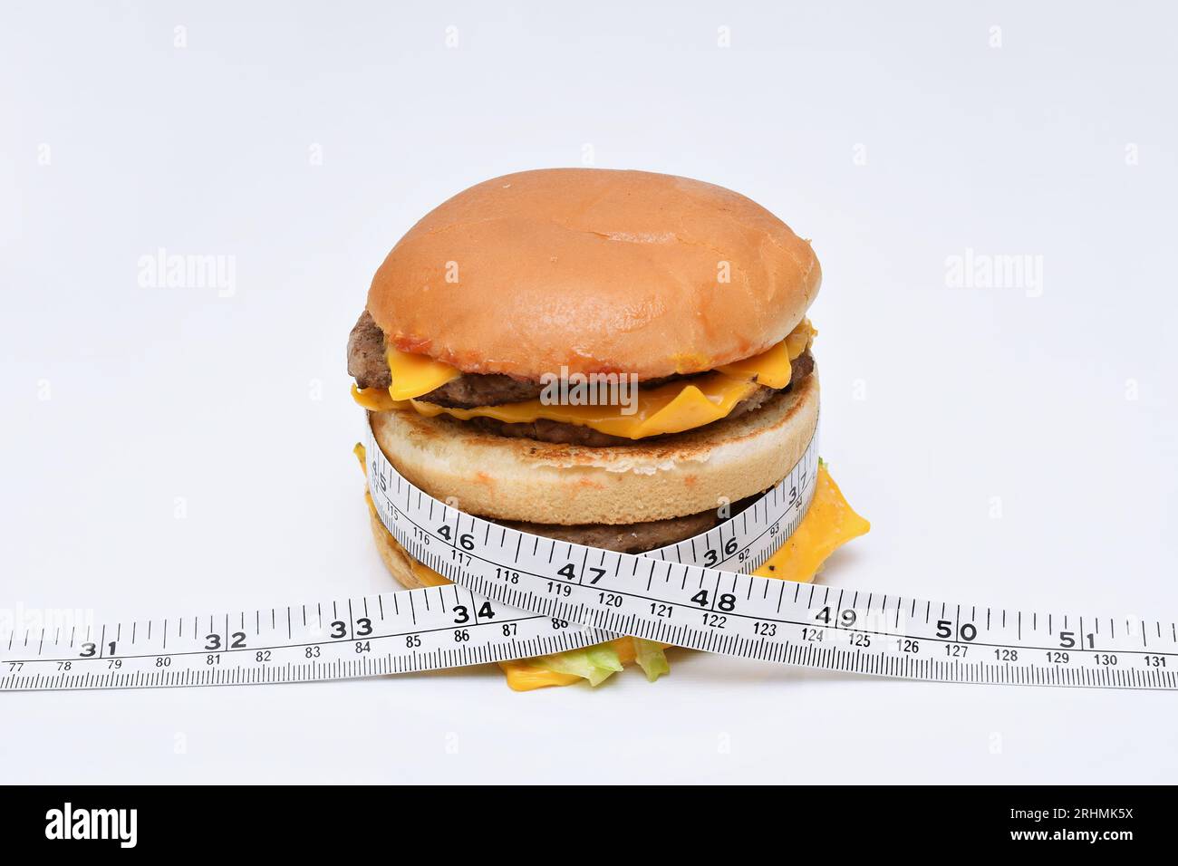Ein Maßband, das um einen fetten, mehrschichtigen Hamburger gewickelt ist, um zu veranschaulichen, wie zu viel Junk Food gesundheitliche Probleme verursachen kann, auf weißem Hintergrund Stockfoto