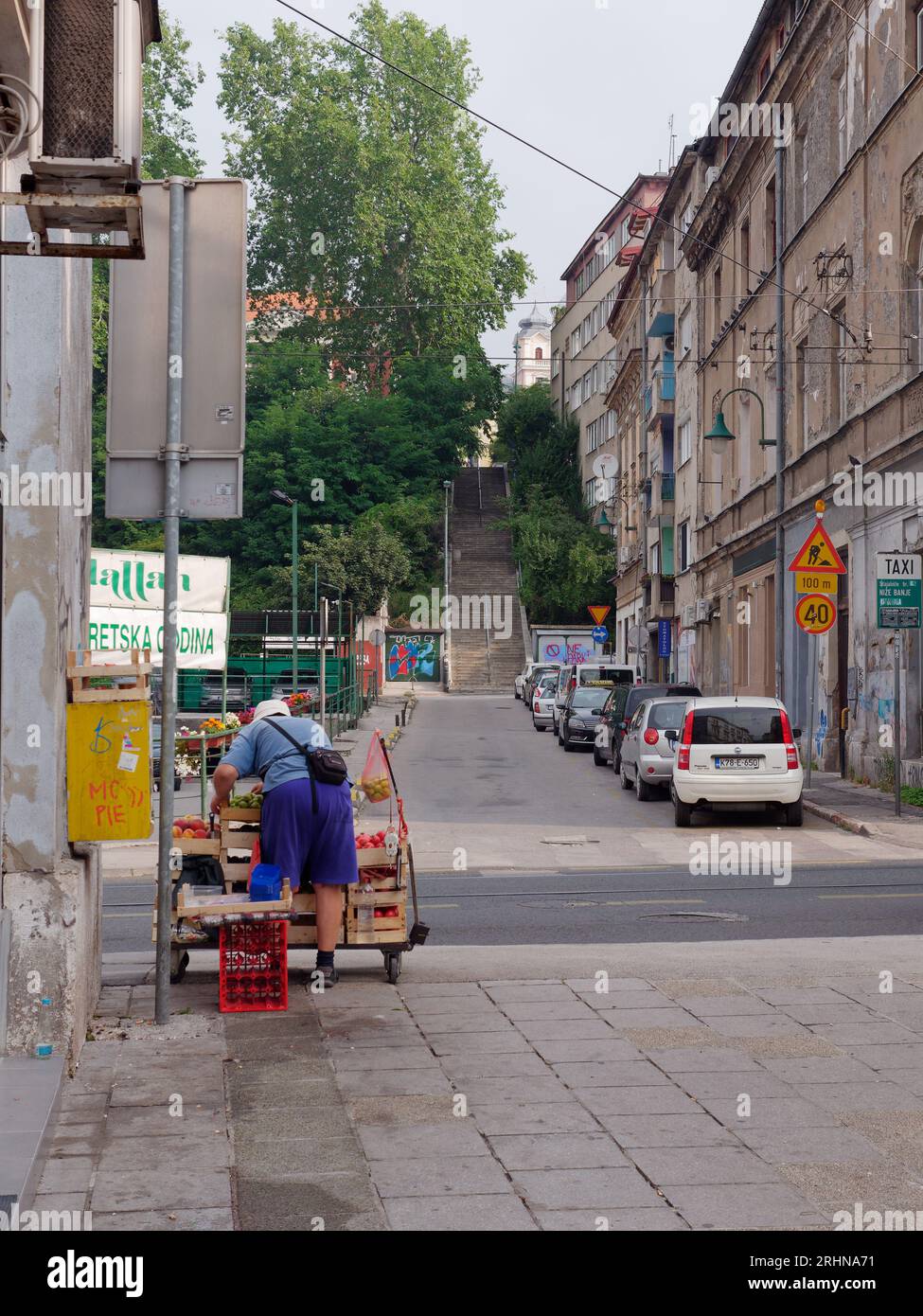 Straße in Sarajevo mit Autos und Stufen, während ein Einheimischer Obst und Gemüse zum Verkauf auf dem Bürgersteig zubereitet, Bosnien und Herzegowina, 18. August 2023. Stockfoto