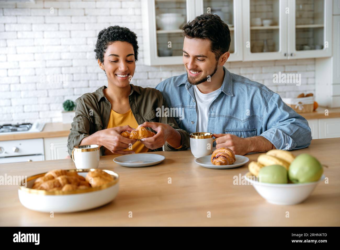 Leckeres Frühstück. Glückliches multirassisches Paar, kaukasischer Mann und afroamerikanische Frau, verbringen Zeit zusammen zu Hause, trinken Kaffee, essen Croissants, unterhalten sich über verschiedene Themen, lächeln Stockfoto