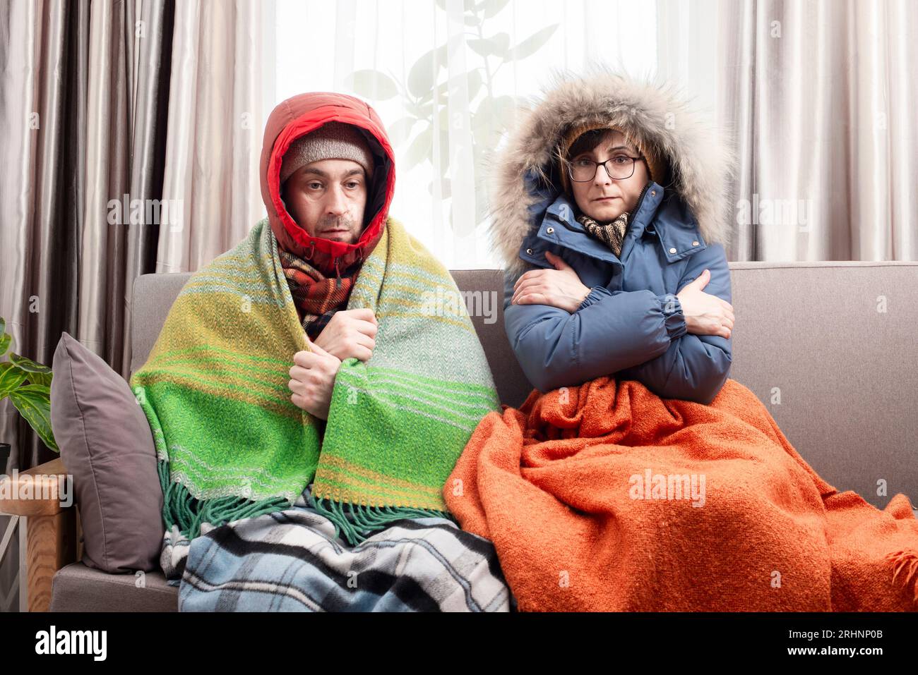 Trauriges Paar mittleren Alters in warmen Kleidern, das auf dem Sofa in ihrem Wohnzimmer kalt wird Stockfoto