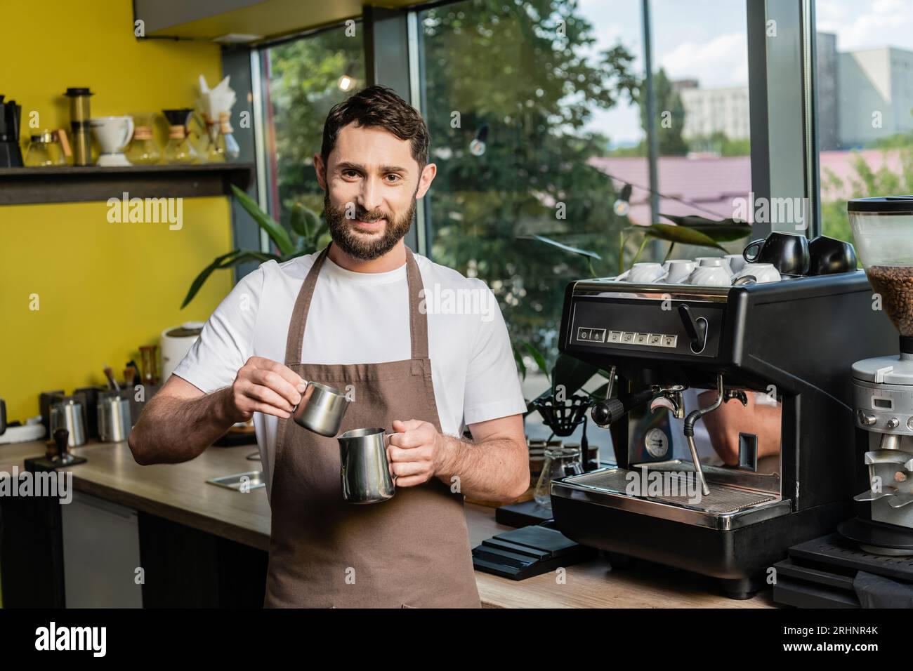 Fröhlicher bärtiger Barista in der Schürze, der in der Nähe einer Kaffeemaschine im Coffee Shop Krüge hält Stockfoto