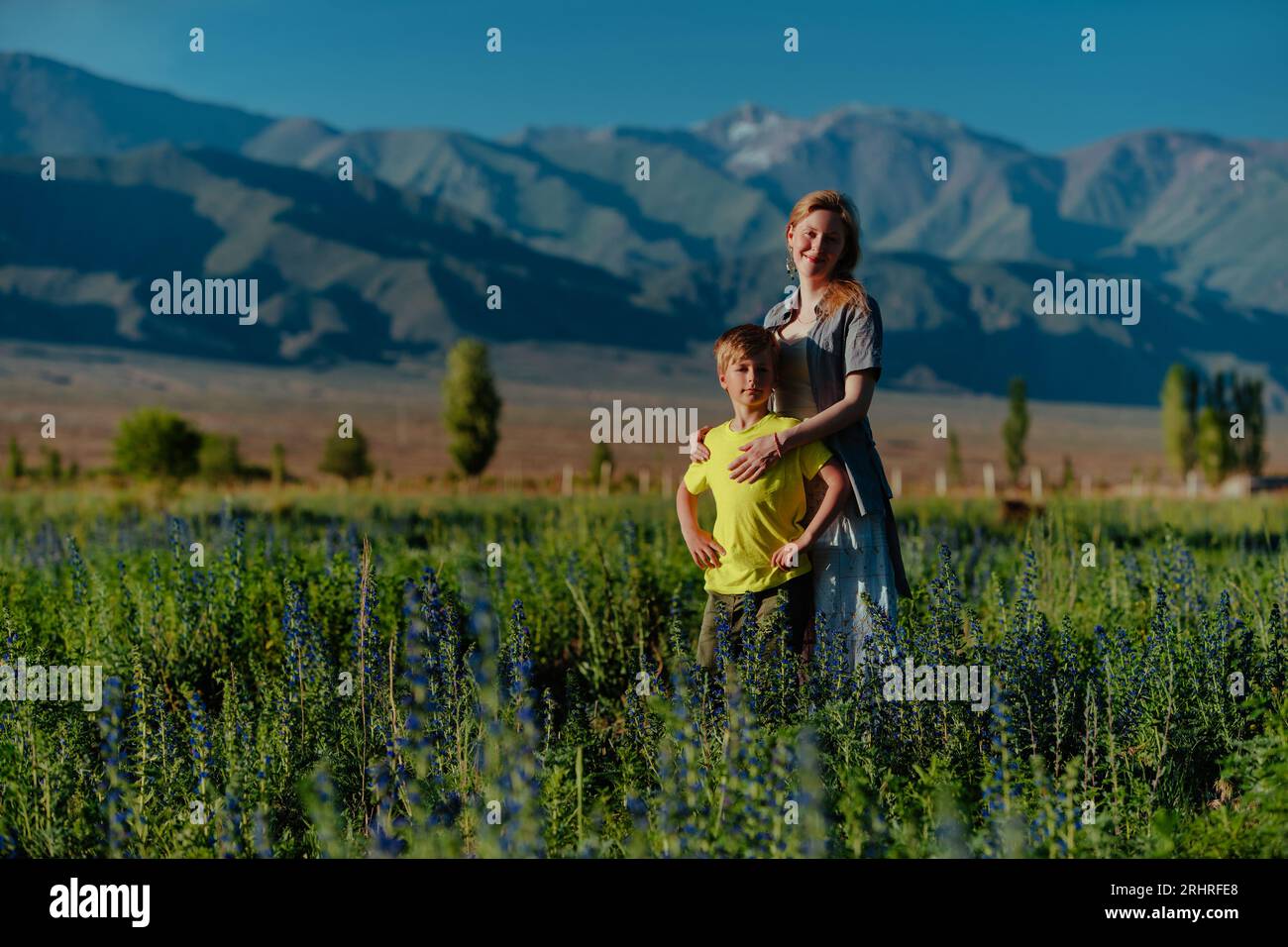 Glückliche junge Frau mit ihrem Sohn, der sich auf einer blühenden Wiese auf dem Hintergrund der Berge posiert Stockfoto