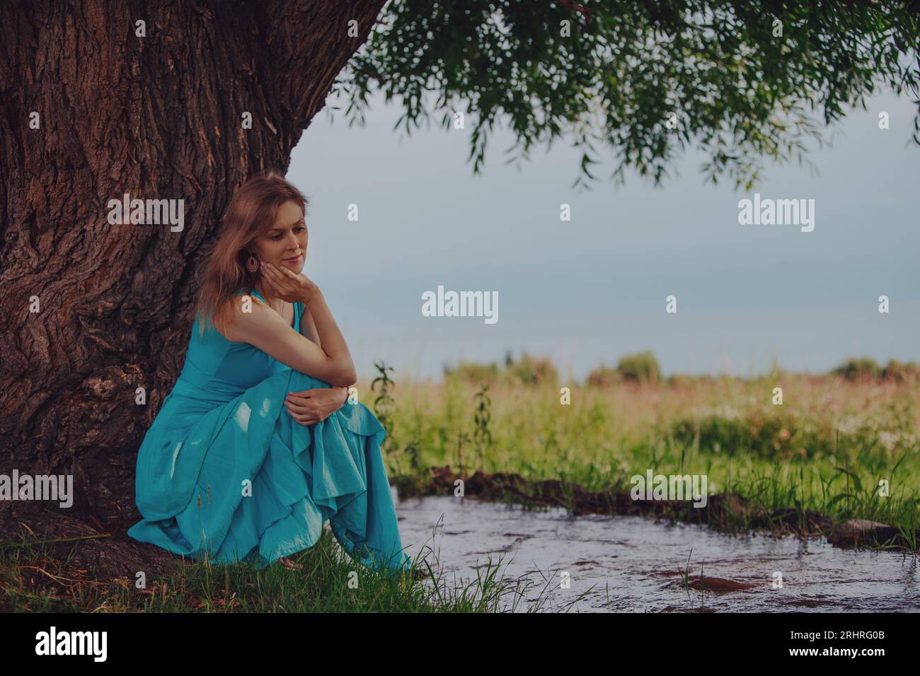 Nachdenkliche elegante Frau in blauem Kleid, die unter einem Baum am Bach sitzt Stockfoto
