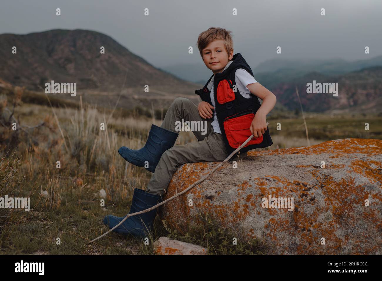 Junge Wanderer mit Stock in einer Weste und Gummistiefeln, die auf dem Stein in den Bergen sitzen Stockfoto