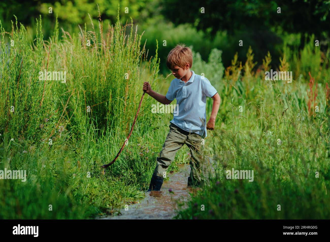 Junge in Gummistiefeln mit Stöcken spaziert an einem flachen Bach entlang Stockfoto
