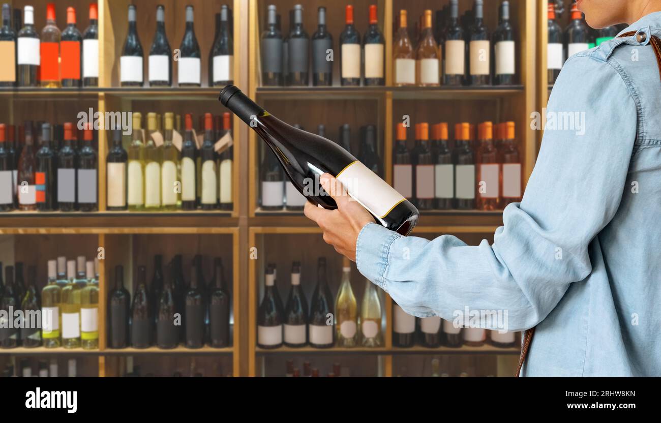Weibliche Person, die eine Rotweinflasche in der Hand vor Regalen hält, mit Weinflaschen im Spirituosengeschäft. Stockfoto
